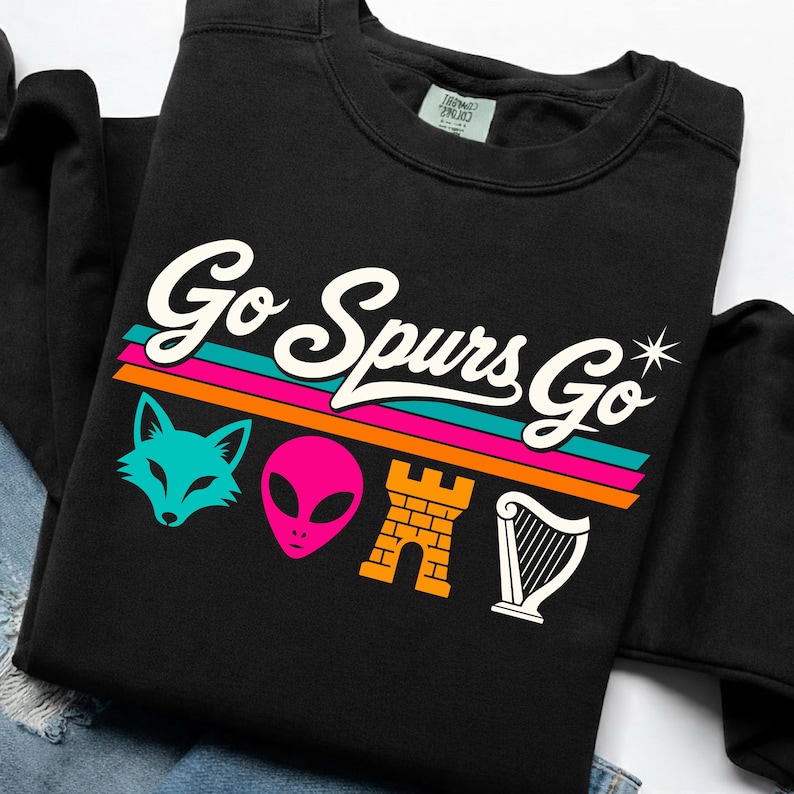K&ouml;nnte beinhalten: Schwarzes Sweatshirt mit dem wei&szlig;en Schriftzug "Go Spurs Go". Darunter befinden sich farbige horizontale Streifen und Cartoon-Grafiken eines Fuchses, eines Aliens, eines Schlosses und einer Harfe. L&auml;ssiger Stil.