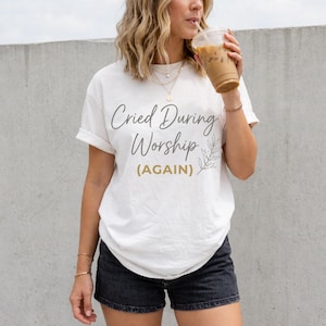 May include: White t-shirt with the text "Cried During Worship (AGAIN)" in a script font. The shirt has rolled-up sleeves and is paired with black shorts. A sprig of leaves is next to the text.