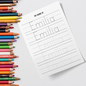 Puede incluir: Una hoja de trabajo imprimible con el texto "MY NAME IS" en la parte superior. El nombre "Emilia" está escrito en líneas punteadas para practicar el trazado. La hoja de trabajo está diseñada para niños que están aprendiendo a escribir su nombre.