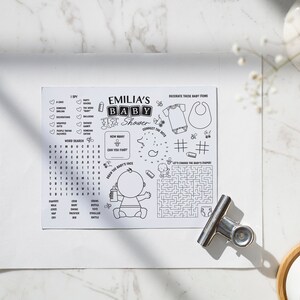 K&ouml;nnte beinhalten: Schwarzwei&szlig;es druckbares Babyparty-Spiel mit Aktivit&auml;ten wie Punkte verbinden, Wortsuche und einem Labyrinth. Das Spiel zeigt ein Baby mit einer Flasche und den Text "Emilia's Baby Shower".