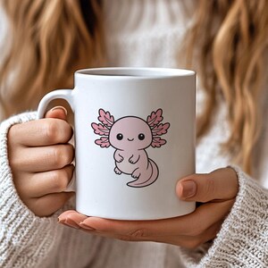 K&ouml;nnte beinhalten: Wei&szlig;e Keramik-Tasse mit einem Cartoon-Axolotl-Design. Der Axolotl ist rosa mit krausen Kiemen und einem l&auml;chelnden Gesichtsausdruck. Die Tasse hat einen Standardgriff. Die Tasse wird von einer Person gehalten, die einen wei&szlig;en Pullover tr&auml;gt.