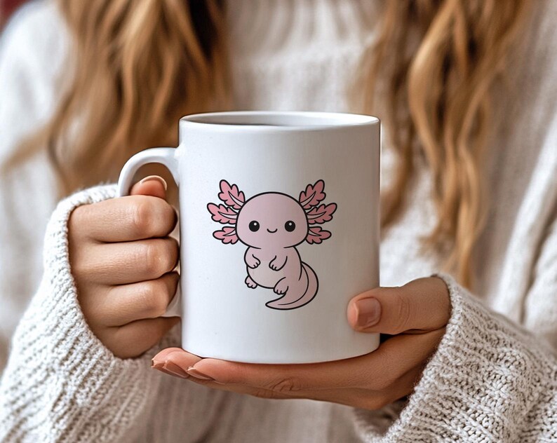 K&ouml;nnte beinhalten: Wei&szlig;e Keramik-Tasse mit einem Cartoon-Axolotl-Design. Der Axolotl ist rosa mit krausen Kiemen und einem l&auml;chelnden Gesichtsausdruck. Die Tasse hat einen Standardgriff. Die Tasse wird von einer Person gehalten, die einen wei&szlig;en Pullover tr&auml;gt.
