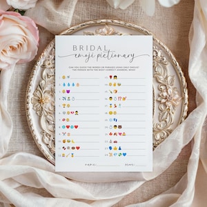 May include: A white Bridal Emoji Pictionary game card with a decorative script font. The card features a series of emoji-based clues for a bridal shower game. The card is on a decorative, gold-rimmed plate, with flowers and fabric in the background.