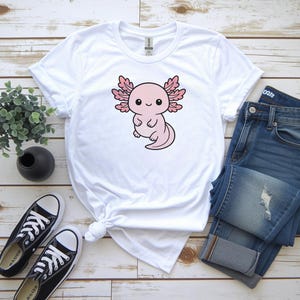 K&ouml;nnte beinhalten: Wei&szlig;es T-Shirt mit einem rosa Cartoon-Axolotl-Design. Das Bild zeigt blaue Jeans und schwarze Leinenschuhe. Der Axolotl ist der Mittelpunkt, mit einem niedlichen Ausdruck. Das T-Shirt ist an der Taille geknotet.