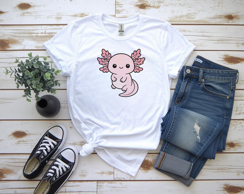 K&ouml;nnte beinhalten: Wei&szlig;es T-Shirt mit einem rosa Cartoon-Axolotl-Design. Das Bild zeigt blaue Jeans und schwarze Leinenschuhe. Der Axolotl ist der Mittelpunkt, mit einem niedlichen Ausdruck. Das T-Shirt ist an der Taille geknotet.
