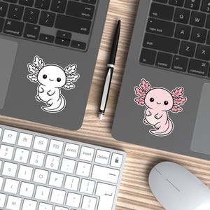 K&ouml;nnte beinhalten: Zwei Laptops mit Axolotl-Aufklebern. Einer ist wei&szlig;, der andere rosa. Ein schwarzer Stift liegt zwischen den Laptops. Eine wei&szlig;e Tastatur und Maus befinden sich im Vordergrund. Die Aufkleber sind im Cartoon-Stil.