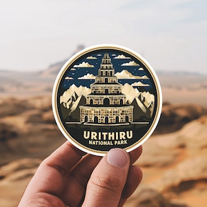 May include: A round sticker with a dark blue background featuring a gold and brown illustration of a tower with a starry night sky and mountains in the background. The text "Urithiru National Park" is printed in gold at the bottom of the sticker.