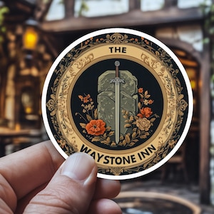 Può includere: Un adesivo rotondo con un disegno di spada nella pietra, con il testo "THE WAYSTONE INN". L'adesivo ha uno sfondo nero con accenti floreali arancioni e dorati. L'adesivo è tenuto in mano.