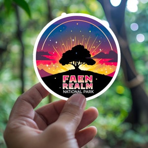 May include: A round sticker with a colorful sunset design featuring a silhouetted tree and the text "FAEN REALM NATIONAL PARK." The sticker has a white border and is held against a blurred green background.