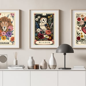 K&ouml;nnte beinhalten: Drei gerahmte Drucke mit floralen Designs und Tarotkarten-Bildern. Der erste Druck zeigt einen L&ouml;wenkopf mit dem Wort "MUT" darunter. Der zweite Druck zeigt eine Eule mit dem Wort "WEISHEIT" darunter. Der dritte Druck zeigt ein Herz mit dem Wort "M&Auml;SSIGKEIT" darunter.