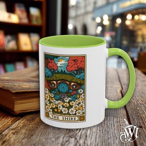May include: White ceramic mug with a green interior and handle. The mug features a colorful illustration of a hobbit hole with the text "THE SHIRE" on the front. The design includes a blue door, flowers, and trees.