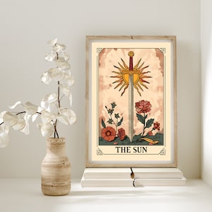 May include: A vintage-style tarot card illustration of The Sun card. The card features a sword piercing a sun with rays, surrounded by flowers. The card is framed in a wooden frame and has a beige background.