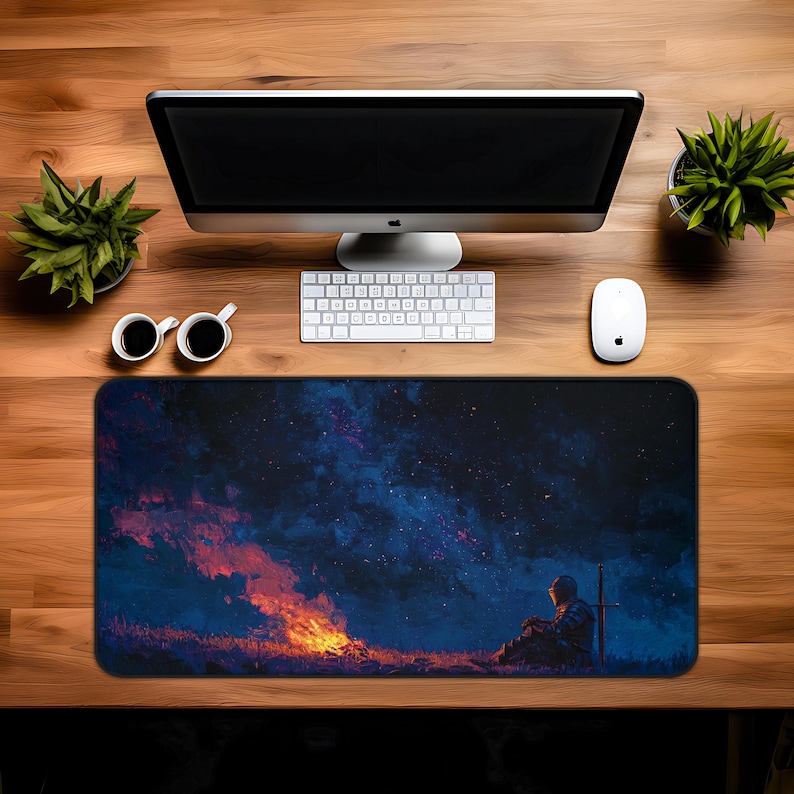 May include: A rectangular mouse pad featuring a digital painting of a knight sitting near a campfire under a starry sky. The pad has a black border and is placed on a wooden desk with a computer, keyboard, mouse, and coffee cups.
