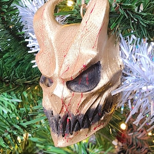 Peut inclure: Une décoration de Noël en forme de crâne démoniaque de couleur dorée avec des accents rouges. L'ornement est suspendu à une branche d'arbre.