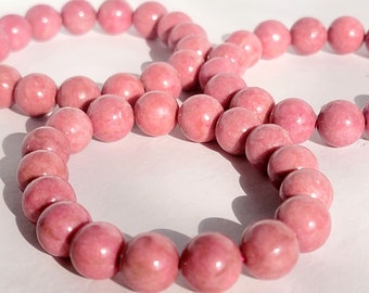 May include: A strand of polished, spherical pink rhodonite beads arranged in a circular pattern. The beads feature natural variations in color, ranging from soft rose to deeper magenta tones, with a smooth, glossy finish and visible internal textures.