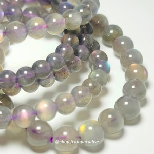 Könnte beinhalten: Ein Perlenarmband aus glatten, runden, durchscheinenden grauen Steinen mit irisierenden violetten, blauen und goldenen Reflexen. Die Perlen sind aufgereiht und der Text @shop.fromparadise ist unten im Bild in Weiß aufgedruckt.