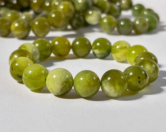May include: A stretch bracelet composed of smooth, round, olive-green gemstone beads with natural variations in color and internal inclusions. A second, matching strand of beads is visible in the background. The text @SHOP.FROMPARADISE is printed at the bottom.