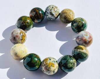 Puede incluir: Una pulsera circular compuesta por doce cuentas esféricas pulidas. Las cuentas presentan una variedad de patrones de piedra natural en tonos verde bosque, crema, marrón y blanco, con algunas mostrando texturas moteadas y otras superficies lisas y brillantes.