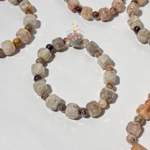 May include: A beaded bracelet featuring faceted, cube-shaped beige and grey stones alternating with small, smooth, dark purple and brown spherical beads. A small logo with the text From Paradise is visible above the center of the bracelet.