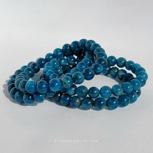 May include: Three blue apatite bead bracelets. Each bracelet is made of round, polished beads. The beads have a deep teal color with variations in tone. The bracelets are arranged in a layered display, showcasing the natural stone's beauty.