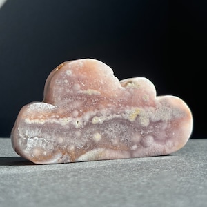 Puede incluir: Una piedra decorativa en forma de nube en tonos rosa, blanco y marrón. La piedra tiene una superficie pulida y estructuras cristalinas visibles. La talla de la nube se encuentra sobre un fondo oscuro.