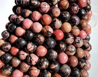 Puede incluir: Una colección de cuentas redondas y pulidas de piedras preciosas en tonos rosa, marrón y negro. Las cuentas tienen patrones únicos y variaciones de color, creando una apariencia natural y texturizada. Es probable que las cuentas estén ensartadas para crear joyas.