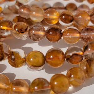 May include: A close-up of a strand of brown and clear gemstone beads. The beads are round and have a smooth, polished finish. The beads are strung on a clear elastic cord.