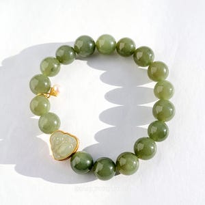 May include: A jade bead bracelet with a gold charm and a small pearl. The bracelet features large, round, olive-green jade beads. The gold charm is in the shape of a small Buddha. The bracelet is on a white surface.