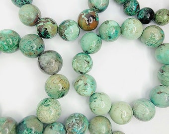May include: A collection of round, polished gemstone beads in varying shades of teal, turquoise, and mint green, featuring natural dark brown and black matrix inclusions. The beads are arranged in a loose, overlapping circular pattern.