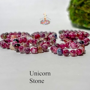 May include: A beaded bracelet made of round, polished stones in shades of magenta, deep pink, and translucent white. The beads are strung together in a circular shape. The text "Unicorn Stone" is printed in black below the bracelet.