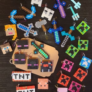 May include: A collection of pixelated keychains made from plastic beads, featuring various shapes including swords, pickaxes, TNT blocks, and character faces in vibrant colors like blue, green, pink, orange, and black, each with a metal ring.