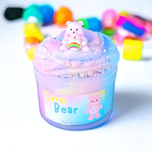 May include: A clear plastic container filled with pink, blue, and white slime. A small pink Care Bear figurine sits on top of the slime. The container has a label that says "Care Bear" and "Fuzzy Slime".