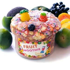 May include: A clear jar of brown slime with a label that says "Fruit Smoothie Fuzzy Slimes". The slime is topped with colorful fruit-shaped candies and sprinkles. The jar is surrounded by various fruits, including limes, avocados, and oranges.