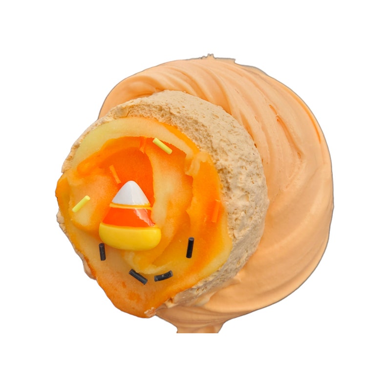 May include: A Halloween-themed cake pop with orange frosting and a candy corn face. The cake pop has a light brown cake base, orange frosting, and is decorated with a candy corn and sprinkles.