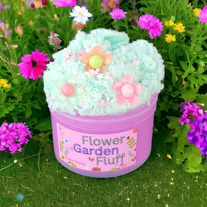 May include: A light green and purple slime with a flower garden theme. The slime is topped with white and green sprinkles, pink and white flowers, and a green and yellow flower. The slime is in a clear container with a pink label that says "Flower Garden Fluff" and "Fuzzy Slimes".