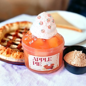 May include: A clear plastic jar filled with a bright orange slime topped with a white fluffy cloud-like topping with pink and blue sprinkles. The jar has a label that says "Apple Pie Fuzzy Slimes".