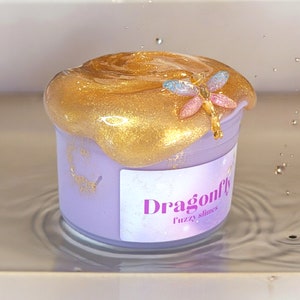 May include: A purple container of gold glitter slime with a dragonfly charm on top. The container has a label that says "Dragonfly Fuzzy Slimes".