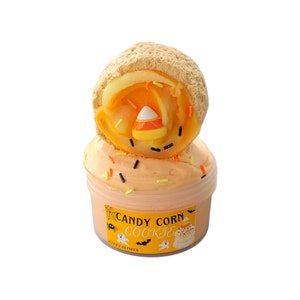 May include: A jar of orange-colored slime topped with a candy corn cookie. The slime is decorated with colorful sprinkles. The jar has a label that reads "Candy Corn Cookie" with Halloween-themed graphics.