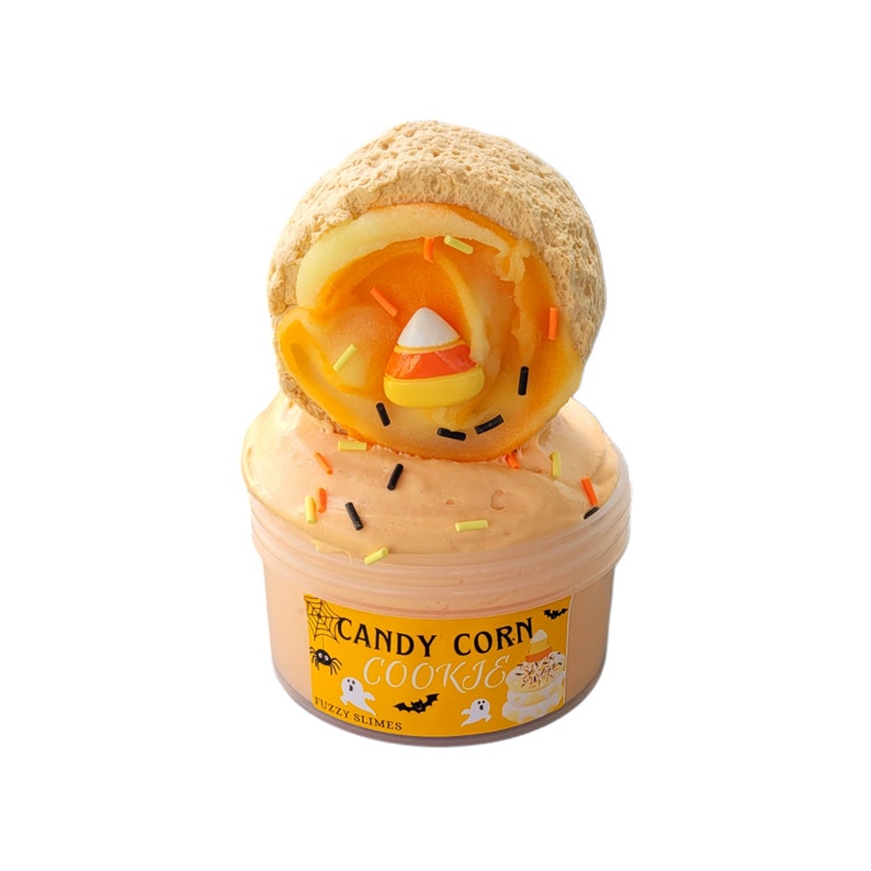 May include: A jar of orange-colored slime topped with a candy corn cookie. The slime is decorated with colorful sprinkles. The jar has a label that reads "Candy Corn Cookie" with Halloween-themed graphics.