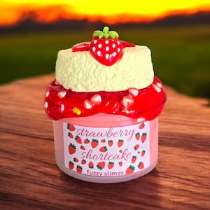 May include: A jar of strawberry shortcake-themed slime. The jar has a light pink base with a label that reads "strawberry shortcake fuzzy slimes." The top is decorated with a yellow cake layer, red strawberry sauce, and a strawberry charm.