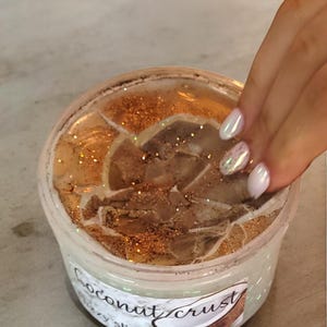 May include: A clear plastic container of "Coconut Crust" slime. The slime has a light brown, textured appearance with gold glitter sprinkled throughout. The container is being touched by a hand with manicured nails.