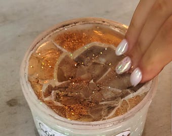 May include: A clear plastic container of "Coconut Crust" slime. The slime has a light brown, textured appearance with gold glitter sprinkled throughout. The container is being touched by a hand with manicured nails.
