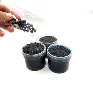 May include: Three clear plastic containers filled with black slime and black beads. A hand is pouring more black beads into one of the containers.