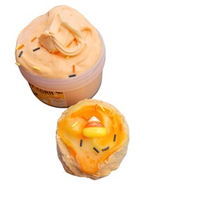 May include: Orange-colored slime in a clear container and a smaller, decorated slime piece. The container is labeled "Candy Corn". The smaller slime is decorated with orange and yellow frosting, candy corn, and sprinkles.
