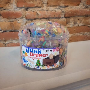 May include: A clear plastic container filled with colorful fuzzy slime charms. The container has a label that reads "Junk Drawer Fuzzy Slimes".