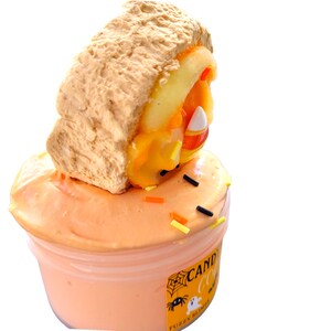 May include: A container of orange slime topped with a faux sandwich. The sandwich has a tan bread-like exterior and a filling of orange and yellow candy corn. Black, orange, and yellow sprinkles are scattered on the slime. The container has a Halloween-themed label.