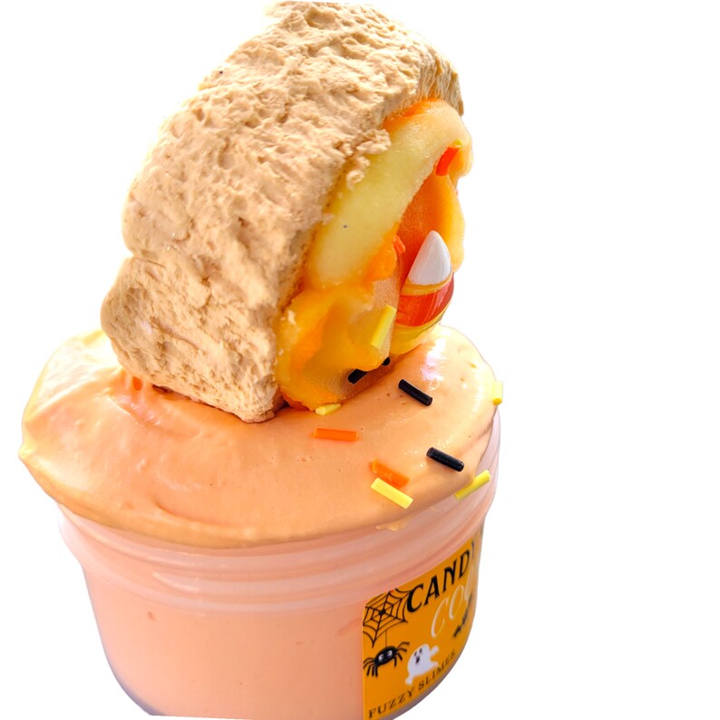 May include: A container of orange slime topped with a faux sandwich. The sandwich has a tan bread-like exterior and a filling of orange and yellow candy corn. Black, orange, and yellow sprinkles are scattered on the slime. The container has a Halloween-themed label.