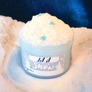 May include: A clear plastic container filled with light blue slime, topped with white, glittery foam and snowflake-shaped confetti. The container has a label that reads "Let it Snow" in cursive script, with a winter forest design. The background is a snowy landscape.