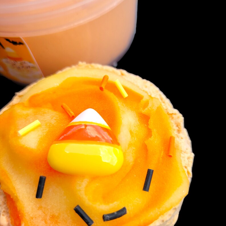 May include: A close-up of a Halloween-themed cupcake with orange frosting, candy corn, and sprinkles. The cupcake has a yellow base and is decorated with black sprinkles to resemble a jack-o'-lantern face. An orange container is in the background.