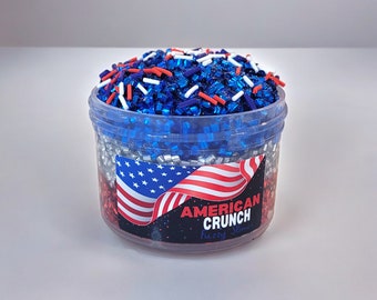 Puede incluir: Un recipiente de plástico transparente lleno de purpurina y espolvorear azul, rojo y blanco. El recipiente tiene una etiqueta que dice "American Crunch Fuzzy Slime".